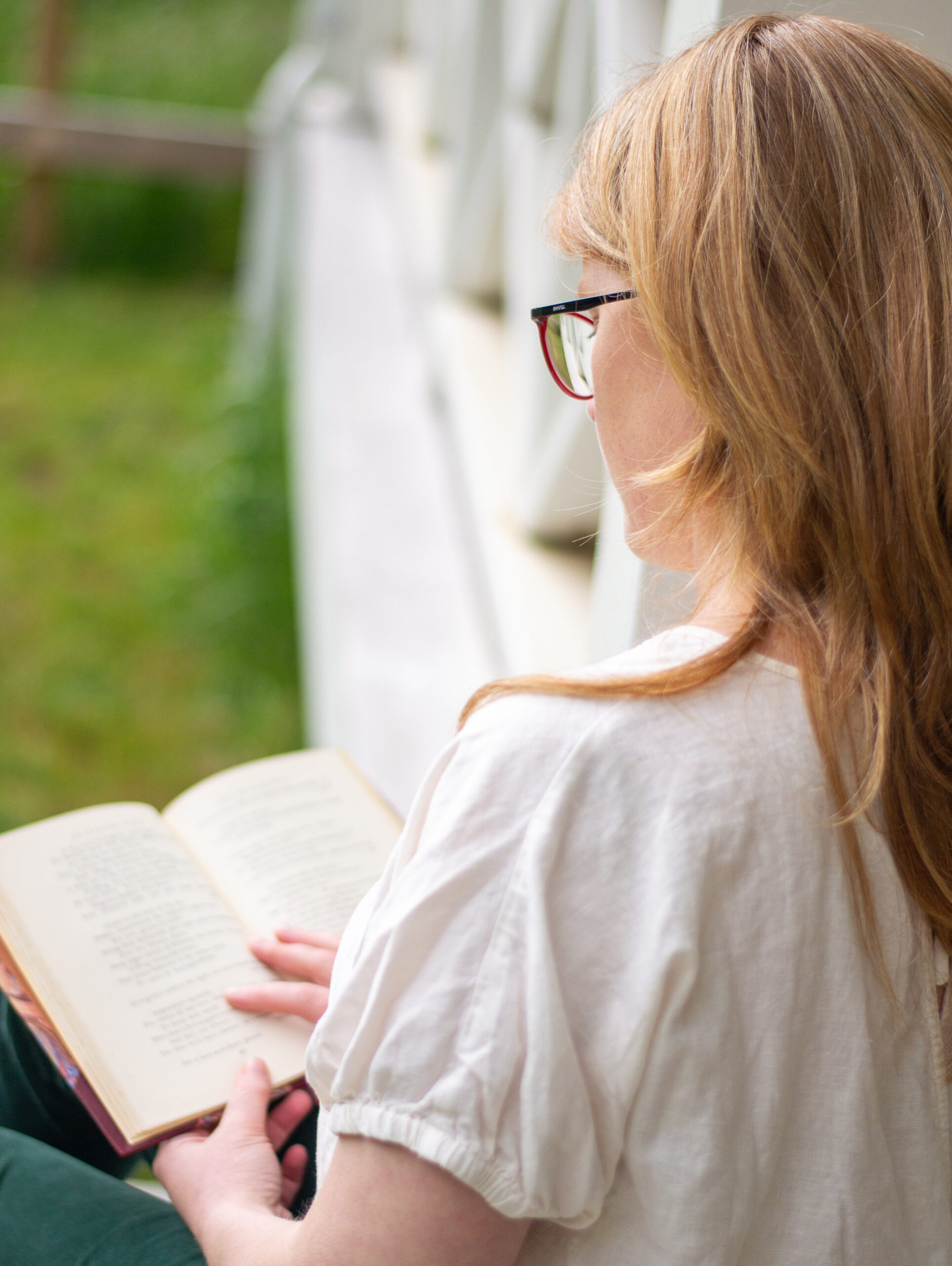 An open book on a table with summer light filtered through a lace curtain. There is a stack of books on the left, and a coffee cup and vase of flowers in the background